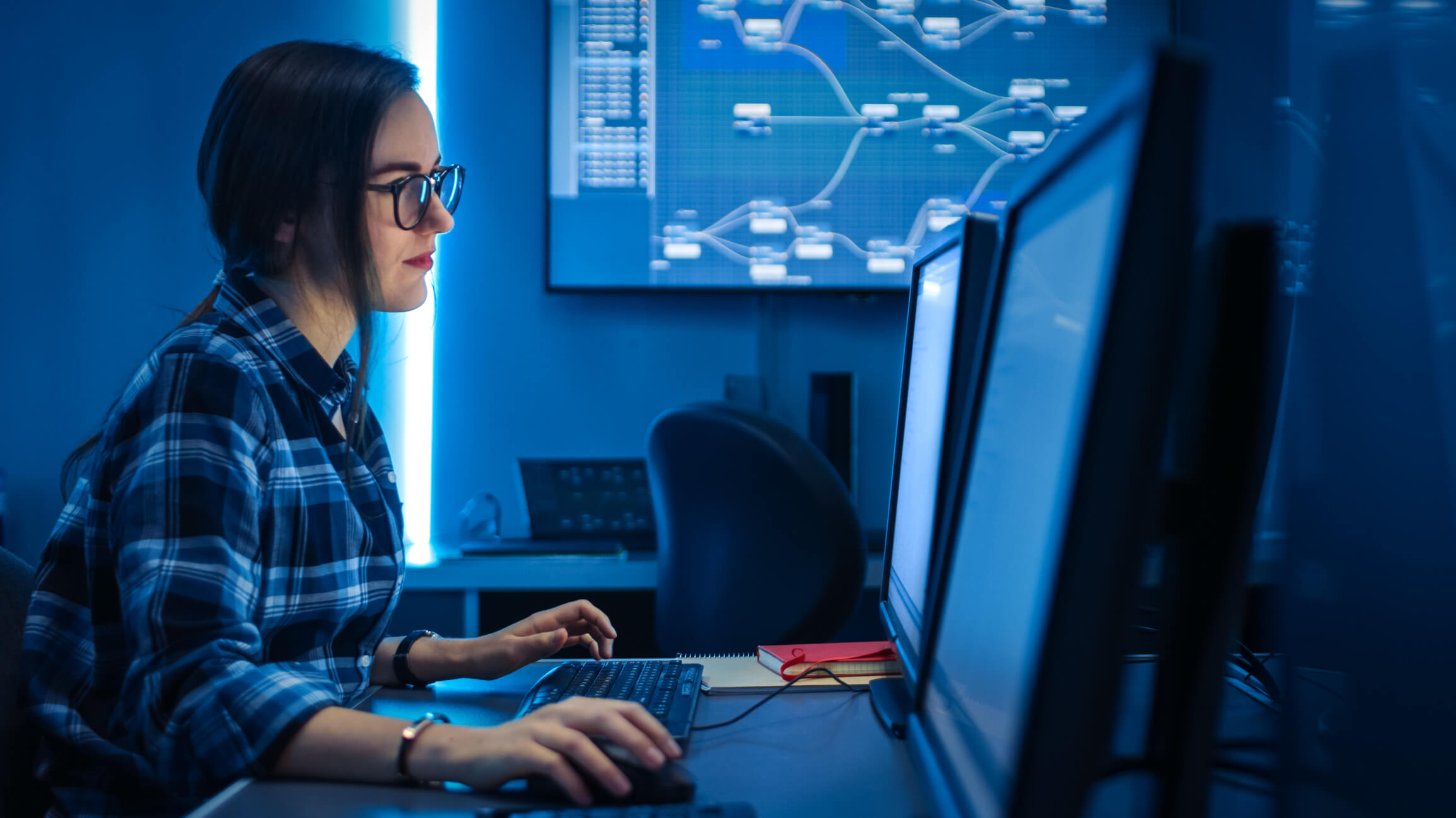 Mulher jovem com a mão no mouse olhando para a tela de um computador em um ambiente com luz azul