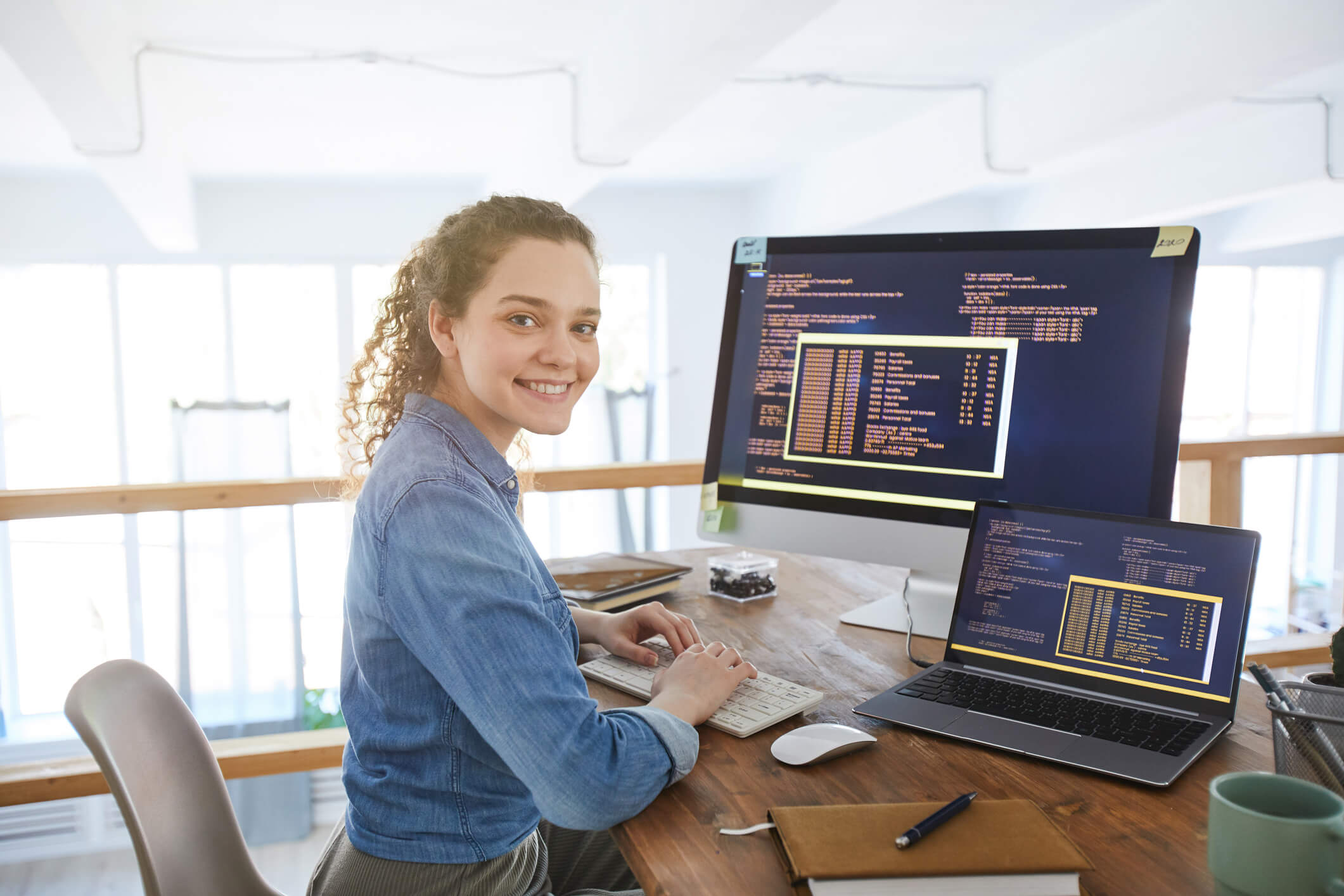 Mulher jovem de cabelos presos olhando para a câmera em frente a uma tela de computador e uma tela de notebook exibindo código-fonte
